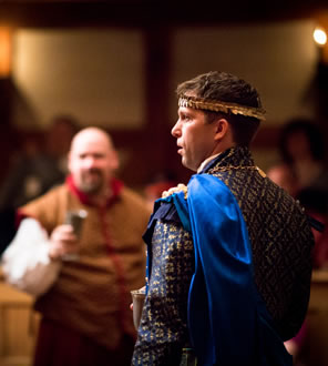 Octavius in blue and gold diamond patterned tunic, blue cape over his left shoulder and gold head band: opposite and out of focus, Enobarbus in brown tunic in red trim and white shirt holds a goblet.